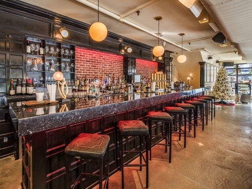 The black bar with red lights under it and sqaure stools in front of a brick wall with liquor