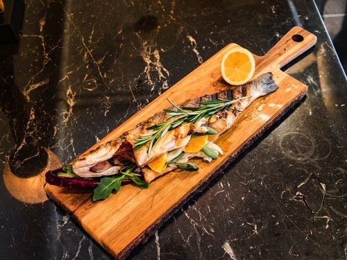 A cooked whole fish sliced open with veggies, lemon and rosemary on a wooden cutting board