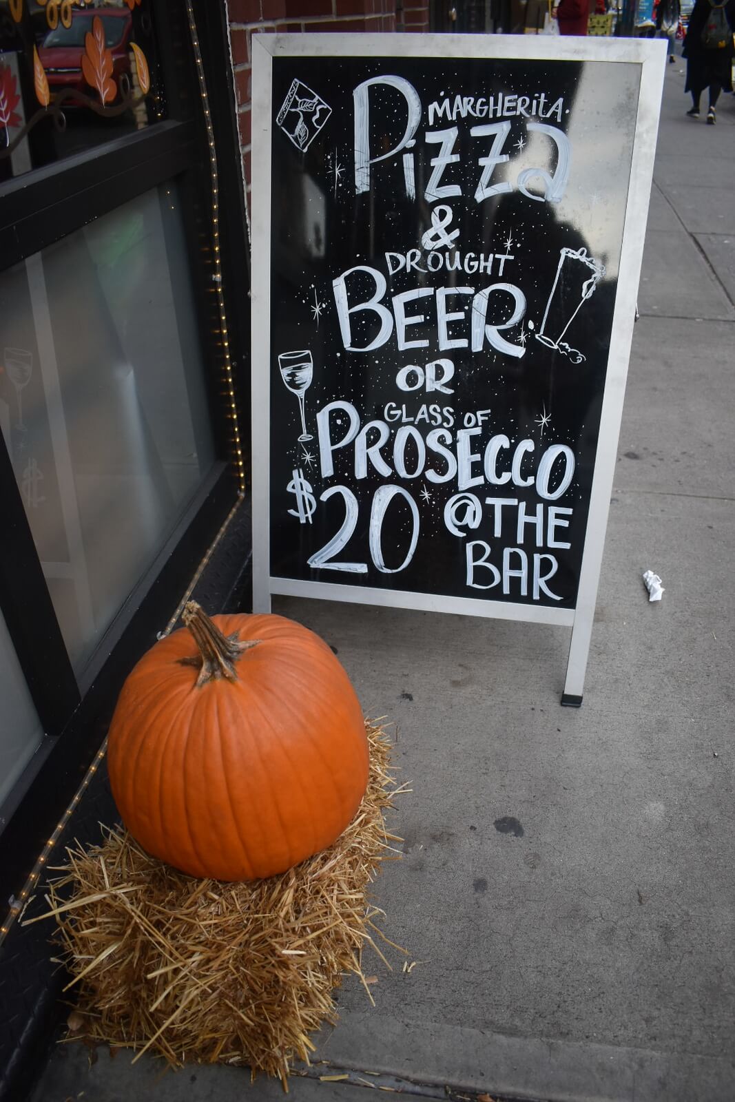 Sidewalk sign that says 'Pizza and Beer or Prosecca $20 at the bar' with a pumpkin in front of it
