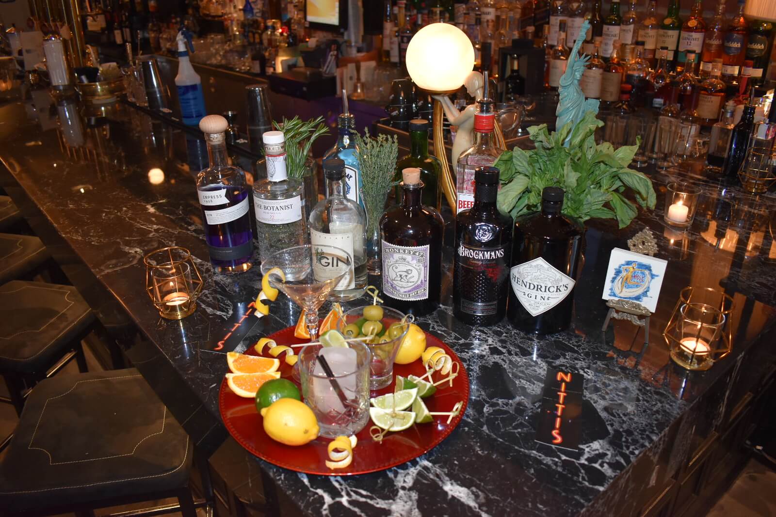 Red plate with a shot glass and citrus slices in front of liquor bottles and herb garnishes on the bar